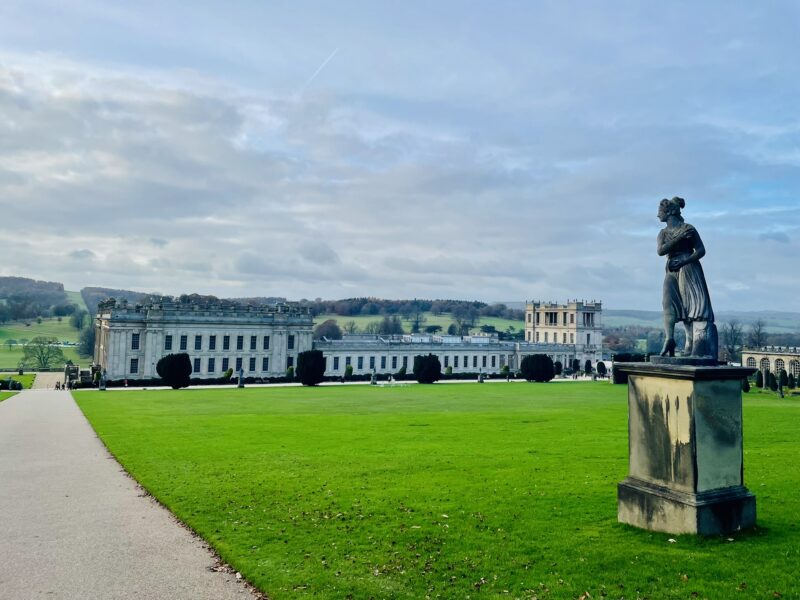 View of Chatsworth House against a green lawn and blue sky. There is a statue to the right hand side.
