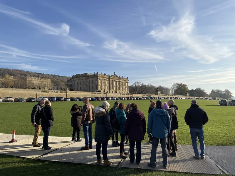 Group of delegates standing on a metal trackway on grass in front of Chatsworth House. Green grass and blue skies make up the background.